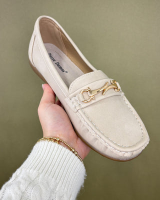 Beige loafer shoe with gold detail held by a hand against a green background