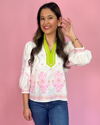 Woman wearing a white blouse with pink floral embroidery and a green neckline against a pink background