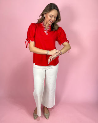Woman wearing a red blouse with white trim and white pants on a pink background