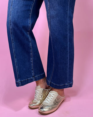 Person wearing blue jeans and gold sneakers on a pink background