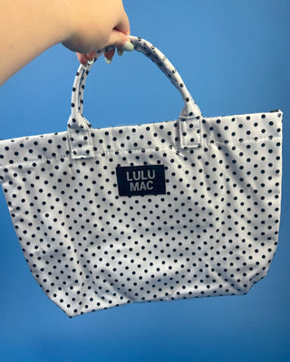A hand holding a white tote bag with black polka dots and a black 'LULU MAC' brand patch on the front, set against a solid blue background.