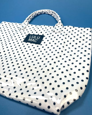 A white tote bag with black polka dots and a black brand label reading 'LULU MAC', photographed on a solid blue background.