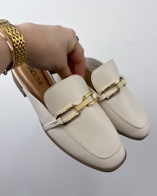 Beige loafers with gold buckles held by a hand on a light gray background
