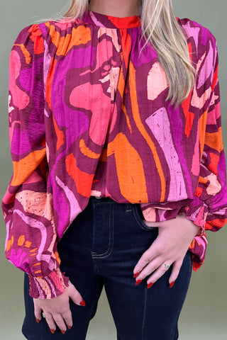 Person wearing a vibrant, patterned blouse with long sleeves in shades of pink, orange, and purple.