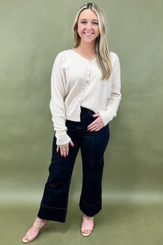 Woman wearing a cream cardigan and dark jeans against a green background