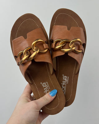 Brown sandals with gold chain detail held by a hand against a gray background
