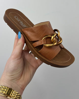 Brown sandal with gold chain held by a hand against a gray background