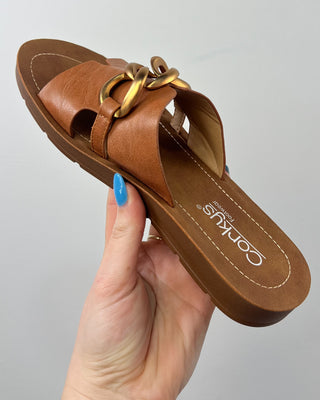 Brown sandal with gold chain held by a hand against a gray background