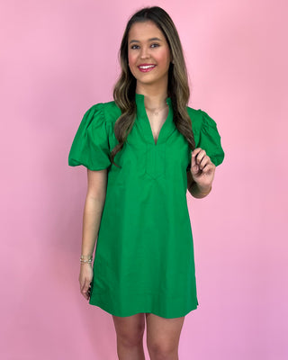 Model wearing an emerald green puff-sleeve mini dress with a split neckline, styled with white heeled sandals against a pink backdrop.