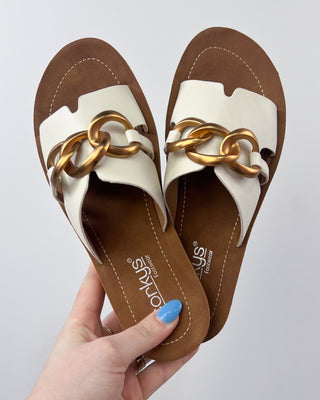 Pair of sandals with gold chain detail held by a hand against a light gray background