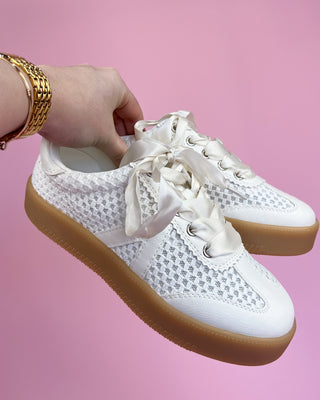 White sneakers with brown soles held by a hand against a pink background