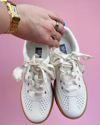 White sneakers with brown soles held by a hand on a pink background