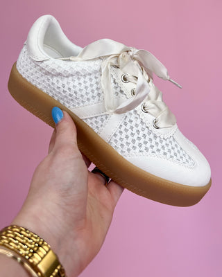 White sneaker with brown sole held by a hand against a pink background