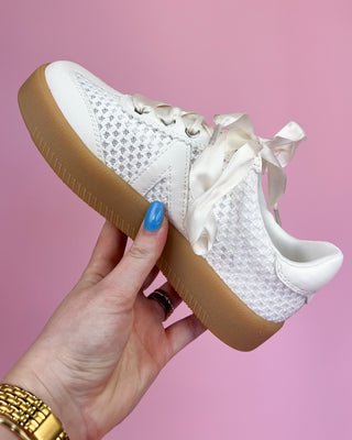 White sneaker with brown sole held by a hand against a pink background