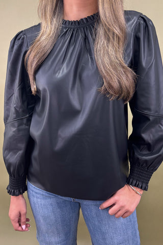 Person wearing a black leather-like blouse with long sleeves and blue jeans on a beige background