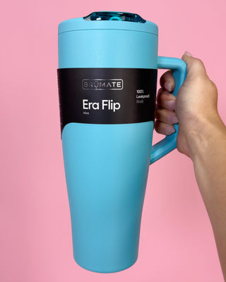 Blue travel mug with 'Brumate Era Flip' branding against a pink background