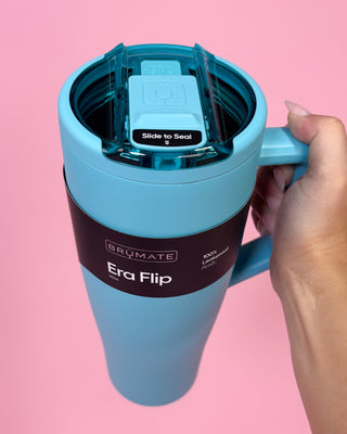 Blue travel mug with handle held by a hand against a pink background