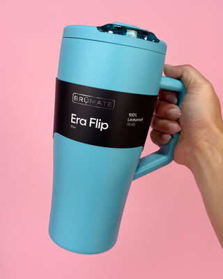 Turquoise mug with black label held by a hand against a pink background