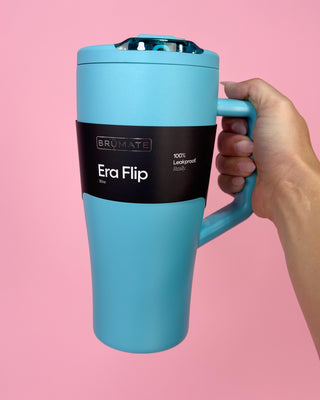 Blue travel mug with black label held by a hand against a pink background