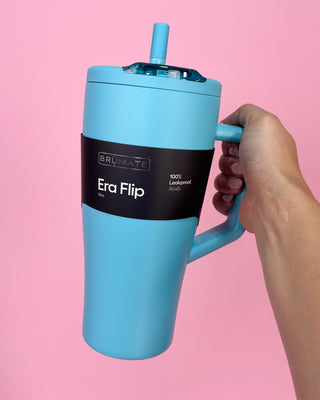 Blue Brumate Era Flip tumbler held by a hand against a pink background