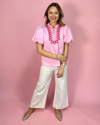 Woman wearing a pink blouse with red embroidery and white pants on a pink background