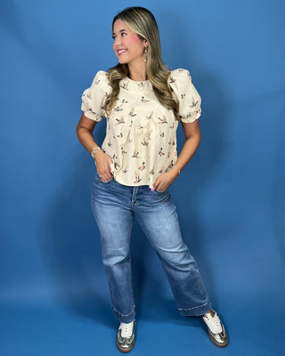 A woman posing in a cream puff-sleeve blouse with a brown and gold bird pattern, paired with blue jeans and white sneakers