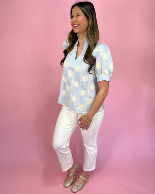 Woman wearing a light blue patterned blouse and white pants against a pink background