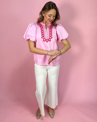Woman wearing a pink blouse with red embroidery and white pants on a pink background