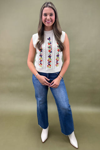 Woman wearing a white sleeveless top with colorful embroidery and blue jeans on a green background