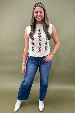 Woman wearing a sleeveless embroidered top and blue jeans against a green background