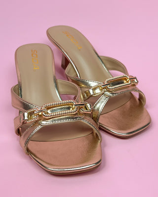 Metallic gold heeled sandals with a square toe, featuring two slim straps and a decorative chain detail across the top, set against a pink background.