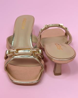 Metallic gold heeled sandals with a square toe, featuring two slim straps and a decorative chain detail across the top, set against a pink background.