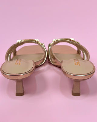 Metallic gold heeled sandals with a square toe, featuring two slim straps and a decorative chain detail across the top, set against a pink background.
