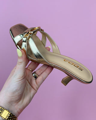 Metallic gold heeled sandals with a square toe, featuring two slim straps and a decorative chain detail across the top, set against a pink background.