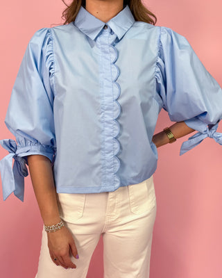 Light blue blouse with ruffled details and tied sleeves worn by a person on a pink background