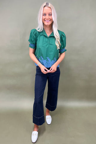 Woman wearing a green blouse with blue floral embroidery and dark jeans on a plain background