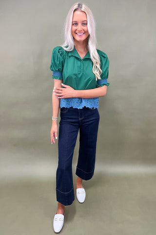 Woman wearing a green blouse with blue trim and dark jeans on a plain background