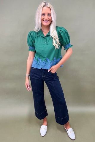 Woman wearing a green blouse with blue lace details and dark blue jeans on a beige background