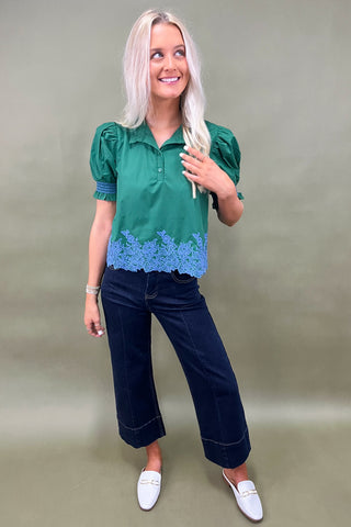 Woman wearing a green blouse with blue lace details and dark blue pants on a beige background
