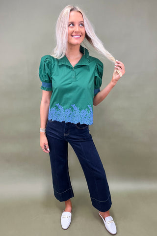 Woman wearing a green blouse with blue lace details and dark pants on a plain background