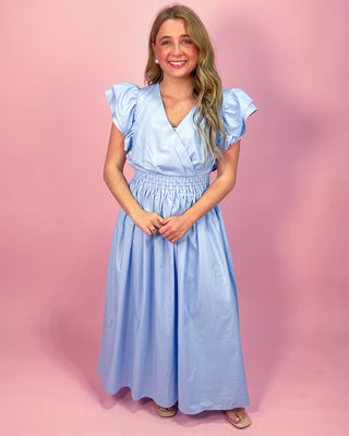Woman wearing a light blue maxi dress with a wrap-style bodice, ruffled cap sleeves, and a smocked waistband, standing against a pink background.