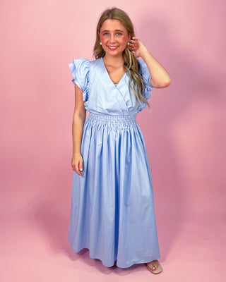 Woman wearing a light blue maxi dress with a wrap-style bodice, ruffled cap sleeves, and a smocked waistband, standing against a pink background.