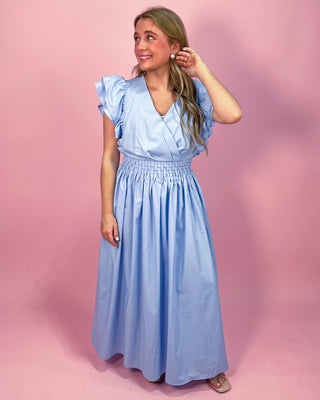 Woman wearing a light blue maxi dress with a wrap-style bodice, ruffled cap sleeves, and a smocked waistband, standing against a pink background.