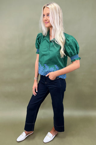 Woman wearing a green blouse with blue floral details and dark pants on a beige background