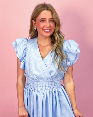 Woman wearing a light blue maxi dress with a wrap-style bodice, ruffled cap sleeves, and a smocked waistband, standing against a pink background.