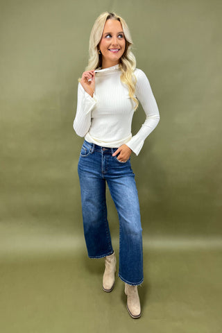 Woman wearing a white long-sleeve top and blue jeans against a green background