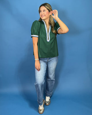 A woman modeling a dark green short-sleeve blouse with white and light blue trim around the V-neck and sleeve cuffs, paired with light-wash blue jeans and metallic sneaker