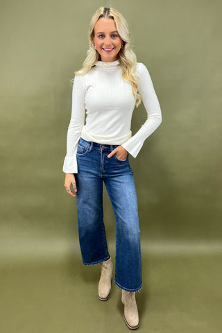 Woman wearing a white long-sleeve top and blue jeans against a green background