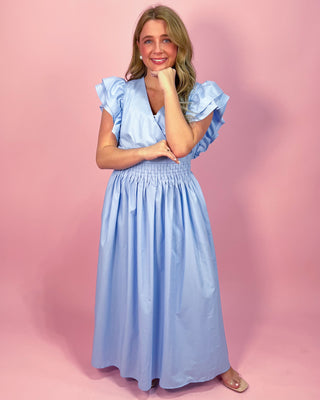 Woman wearing a light blue maxi dress with a wrap-style bodice, ruffled cap sleeves, and a smocked waistband, standing against a pink background.