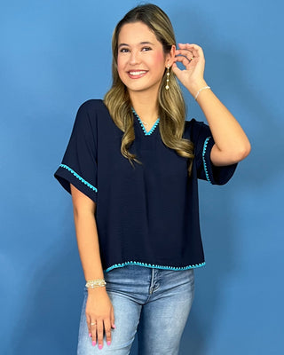 A woman modeling a navy blue short-sleeve top with turquoise trim on the V-neck and sleeve cuffs, paired with light-wash blue jeans